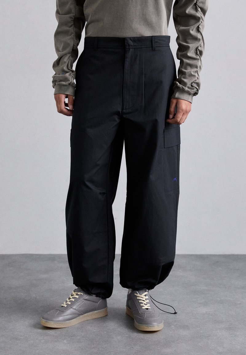 Pantaloni cargo neri con vestibilità rilassata, dotati di tasche laterali e un logo blu discreto. Realizzati in tessuto morbido, abbinati a sneaker grigie.