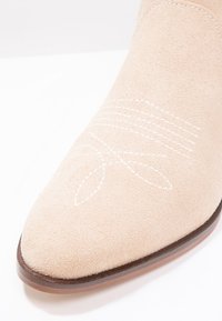 Botín de ante beige hasta el tobillo con punta afilada, que presenta costuras decorativas blancas y una suela marrón. Textura suave con acentos sutiles.