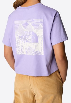 Personne portant un t-shirt court lavande avec un motif de montagne et forêt blanc et le texte "The North Face" au dos, associé à un pantalon en velours côtelé beige.