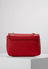 Borsa sintetica rossa con forma strutturata, accenti in catena dorata e chiusura a pattina. Texture liscia con dettagli di cucitura tono su tono.