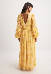 Pitkä keltainen kukkamekko kevyestä kankaasta, jossa on röyhelöhihoja ja syvä V-muotoinen selkäosa, joka kiinnitetään nauhalla.