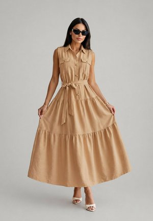 Femme portant une robe maxi beige sans manches boutonnée avec ceinture, tenant les côtés de la jupe, accessoirisée de lunettes de soleil noires et de sandales blanches à talons.