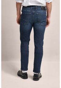 Ciemnoniebieskie dżinsy o slim fit, z dwoma tylnymi kieszeniami i skórzaną naszywką z logo na pasku. Trzymane rękami po bokach.