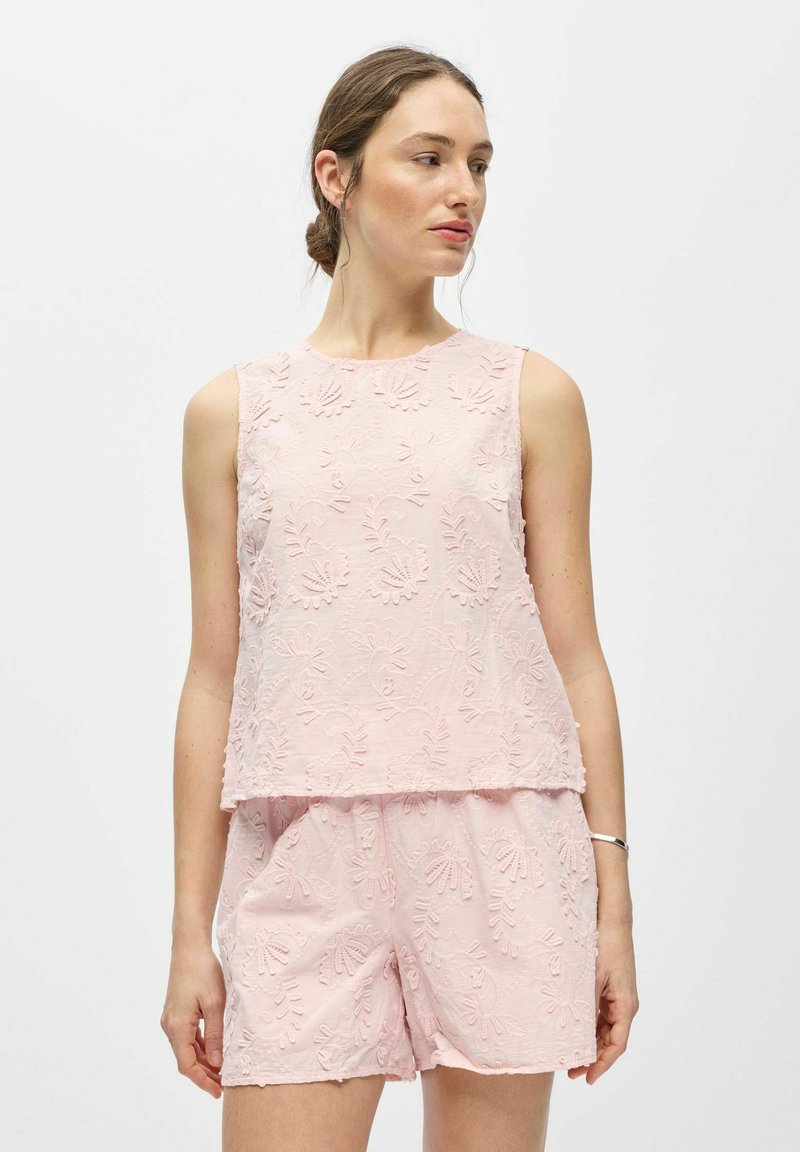 Femme portant un haut sans manches rose pâle à texture florale et un short assorti, debout sur un fond blanc uni.