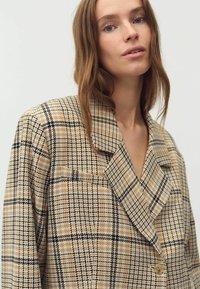 Beige kariertes Blazer mit schwarz-orangefarbenen Akzenten, ausgestattet mit einer Reverskragen, doppelreihigem Design und strukturiertem Stoff.