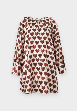 Kvinde iført en hvid kjole til knæet dækket med røde og brune hjerte-mønstre, kombineret med sorte spidse hæle.