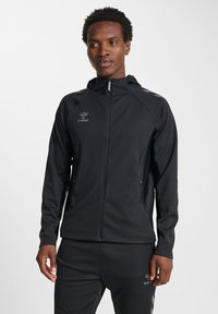 Zwarte sportjas met ritssluiting aan de voorkant, capuchon en reflecterende accenten. Heeft een soepel stof en zijzakken voor opberging.