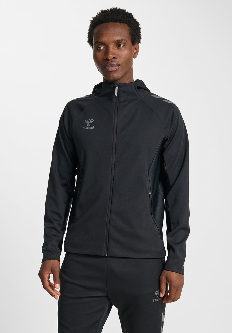 Zwarte sportjas met ritssluiting aan de voorkant, capuchon en reflecterende accenten. Heeft een soepel stof en zijzakken voor opberging.