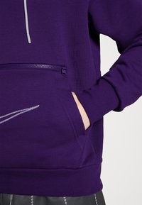 Personne portant un sweat à capuche violet avec une poche zippée à l'avant et la main dans la poche latérale, montrant une partie de la manche et du cordon.