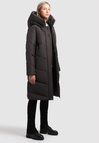 Ung kvinna som står, iklädd en lång mörkbrun vadderad jacka med stor huva, svarta byxor och svarta kraftiga stövlar mot en enfärgad bakgrund.