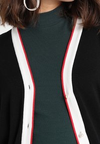 Svart kavaj med vit och röd trim. Inkluderar en ribbad grön topp under. Knapparna har en dekorativ pärlaccents.