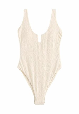 Kremowy jednoczęściowy kostium kąpielowy z teksturowanym wzorem, głębokim dekoltem w kształcie łodzi i wysokim krojem nogawek. Ma wycięcie w kształcie klucza z przodu.