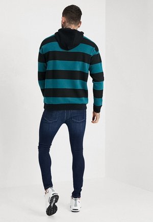 Mann trägt schwarzen und türkis gestreiften Hoodie, dunkle enge Jeans und weiße Sneaker, steht mit dem Rücken zur Kamera.