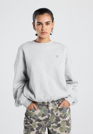 Vrouw met kort, donker haar draagt een lichtgrijs sweatshirt en camouflagebroek met haar handen in de zakken tegen een effen achtergrond.