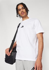 Λευκό βαμβακερό μπλουζάκι με κοντά μανίκια και μαύρο λογότυπο Adidas. Το μοντέλο κρατάει μια μαύρη τσάντα crossbody με υφή και ρυθμιζόμενο λουρί.