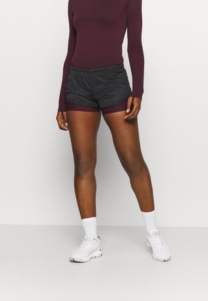 Kvinde iført en bordeauxfarvet langærmet top og shorts med sorte sportsshorts lagdelt over bordeauxfarvede shorts, hvide sokker og hvide sneakers.