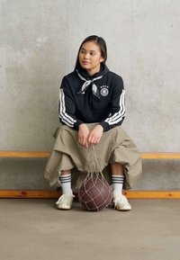 Sudadera negra con rayas blancas, pantalones caqui de gran tamaño y zapatillas blancas. Sosteniendo un balón de baloncesto en una red, sentado en un banco de madera contra una pared de hormigón.