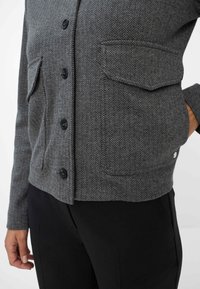 Giacca a spina di pesce grigia con bottoni neri e tasche con pattina, indossata con pantaloni neri, mano appoggiata sui fianchi.