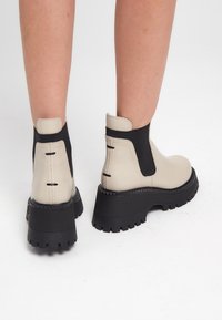 Botas de tobillo de cuero beige con lados elásticos negros, suelas de goma negra gruesa y acentos texturizados. Presentan costuras reforzadas y tiradores.
