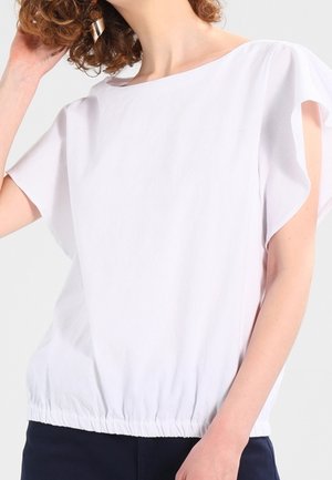 Frau trägt eine weiße kurzärmelige Bluse mit gerafftem elastischem Saum und dunkler Hose, berührt mit einer Hand ihr lockiges Haar.