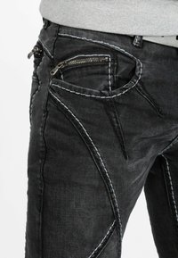 Zwarte denim jeans met opvallende grijze stiksels, schuine zakken en ritsafsluitingen. De stof lijkt lichtelijk versleten met een gladde textuur.