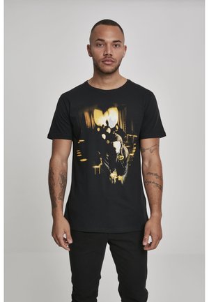Homme portant un t-shirt noir avec un motif graphique abstrait jaune et blanc, debout devant un fond gris clair uni.