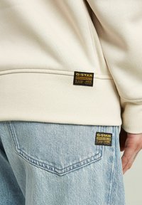 Beige sweatshirt and light blue denim jeans with black and yellow G-Star RAW labels on waistband and back pocket, visible hand by side.