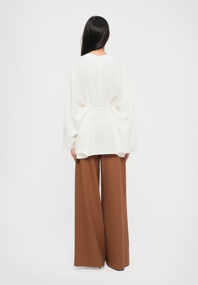 Woman standing facing away, wearing a loose white blouse with batwing sleeves and wide brown pants against a plain background.