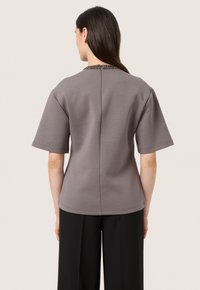Top grigio a maniche corte con scollo a girocollo e un colletto texturizzato con perline. Il design presenta una linea dritta e una chiusura con zip sul retro.