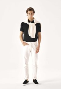 Camisa negra de manga corta, vaqueros blancos con puños doblados y zapatos negros sin cordones. Un suéter de color claro está colocado sobre los hombros.