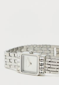 Ezüst fém óra négyzet alakú számlappal, fehér óra számlappal és kristály díszítéssel a kereten. Texturált lánc karkötővel és fényes felülettel rendelkezik.