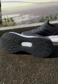 Zapatilla deportiva negra con una suela de goma texturizada, que presenta un acento blanco a lo largo de la mediasuela y un diseño superior liso.