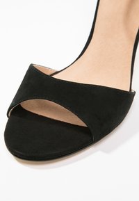 Gros plan d'une sandale ouverte en daim noir avec une large bride avant et une semelle intérieure beige sur fond blanc.