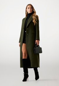Palton din lână de culoare verde măsliniu, cu un croi lung, rever ascuțit și două butoane. Pulover negru cu guler înalt, cizme înalte și o geantă mică neagră.