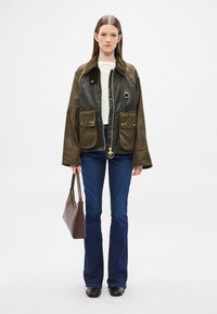 Olive green jacket with black leather front, large pockets, and gold hardware. Paired with flared blue jeans and a brown handbag.