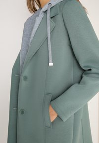 Personne portant un manteau vert par-dessus un sweat à capuche gris zippé, la main dans la poche du manteau, montrant les boutons du manteau et les cordons du sweat à capuche avec du texte.