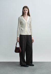 Nő krémszínű és fekete csíkos, gombos kardigánban, széles fekete nadrágban, fekete cipőben, sötétvörös táskát tartva, szürke padlón áll.