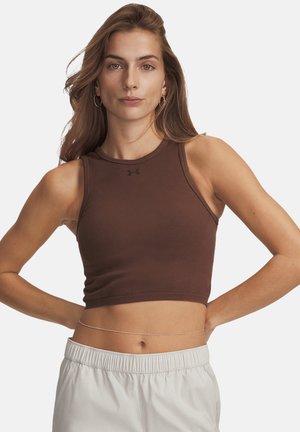 Mujer con cabello largo que lleva pendientes de aro, un top corto marrón sin mangas, pantalones blancos y una cadena fina en la cintura, posando con las manos en las caderas.