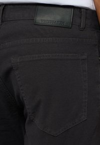 Pantaloni in denim nero con una tasca posteriore e un'etichetta in pelle impressa con "TRUSSARDI". Il tessuto ha una consistenza liscia e cuciture sottili.