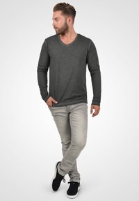 Cămașă cu mânecă lungă, gri închis, cu decolteu în V, asortată cu blugi gri deschis și adidași negri, prezentată pe un model masculin într-un fundal neutru.