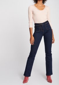 Jeans de tiro alto de color azul oscuro con pierna acampanada, que cuentan con cuatro bolsillos y costuras en contraste, combinados con una blusa de manga larga acanalada de color rosa claro.
