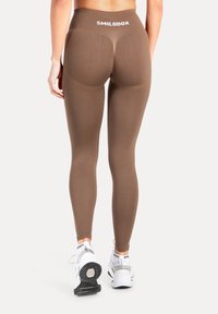 Braune Leggings mit hoher Taille, strukturiertem Finish und betonten Nahtdetails, kombiniert mit weißen Sportschuhen.