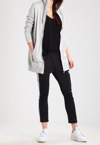 Donna che indossa una felpa grigia con zip, un top senza maniche nero, pantaloni sportivi Adidas neri con strisce bianche e sneakers bianche, in piedi con una mano nella tasca.