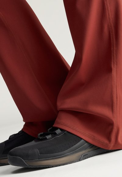 Billedet viser sorte atletiksk shoes med teksturerede såler sammen med rustfarvede bukser, der dækker anklerne.