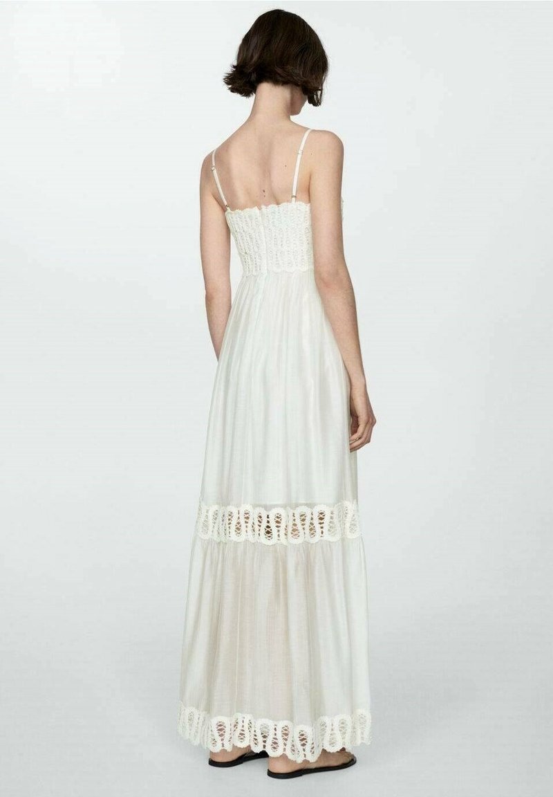 Femme aux cheveux bruns courts portant une longue robe blanche sans manches avec des détails en dentelle dans le dos, debout devant un fond clair uni.