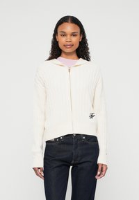 Kremowy hoodie z dzianiny warkoczowej z przednim zamkiem, z ribbowanymi mankietami i dołem; posiada małe haftowane logo po lewej stronie. Ciemne dżinsy na dole.