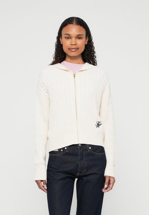 Creme kabelstrikket hoodie med frontlynlås, ribbede ærmer og kant; har et lille broderet logo på venstre side. Mørke jeans nedenfor.