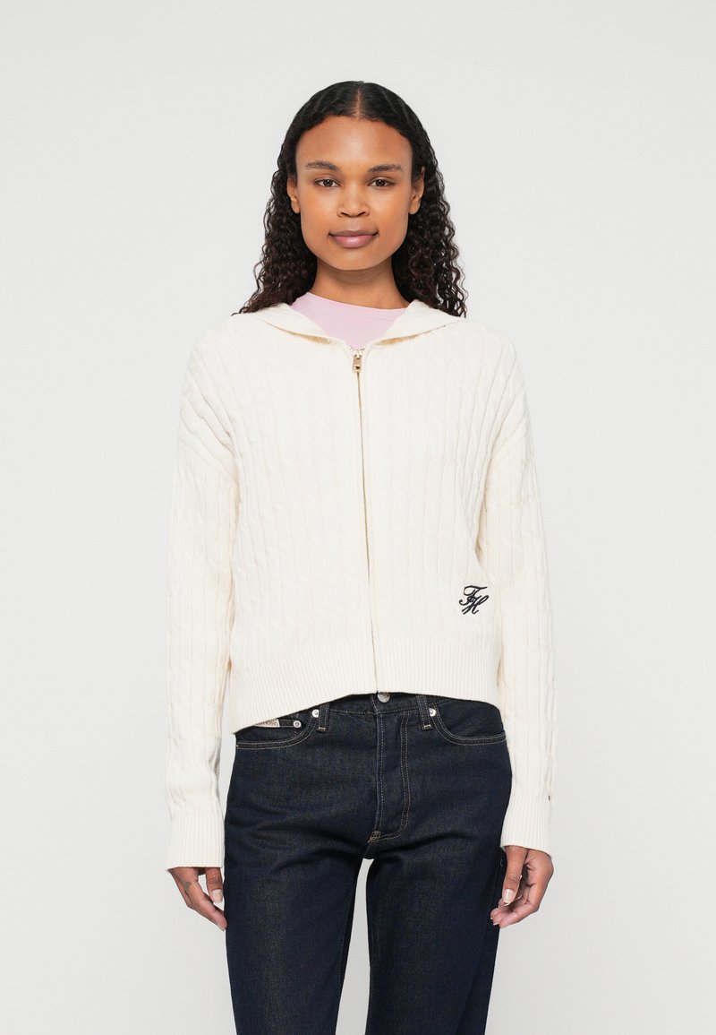 Kremowy hoodie z dzianiny warkoczowej z przednim zamkiem, z ribbowanymi mankietami i dołem; posiada małe haftowane logo po lewej stronie. Ciemne dżinsy na dole.