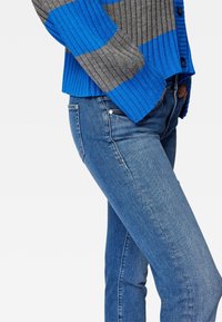 Le cardigan en maille présente des rayures grises et bleues, une texture côtelée, une fermeture à boutons, accompagné d'un jean slim bleu en denim délavé moyen.