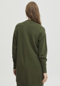 Pull long, oversized de couleur vert foncé, fabriqué en maille douce. Présente une coupe décontractée et des poignets et un ourlet côtelés.